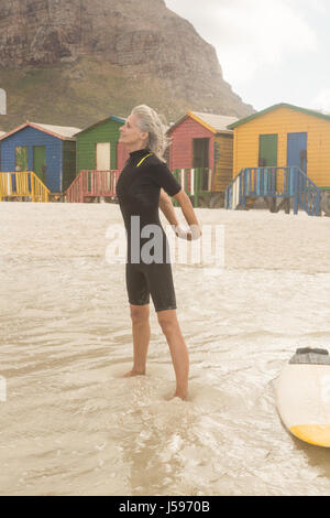 Ältere Frau ausdehnen, während sie von Surfbrett stehend gegen Hütten Stockfoto