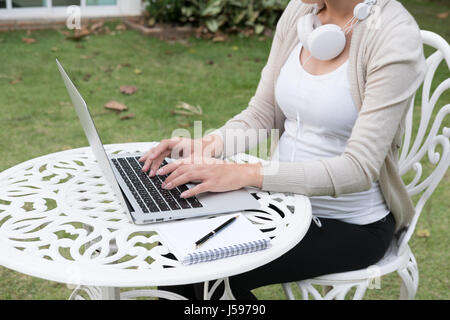 Junge asiatische Frau auf Stuhl sitzend mit Kopfhörer mit Laptop-Computer. Schöne weibliche werdende Kind die Erholung im Garten vor ihrem Haus Stockfoto