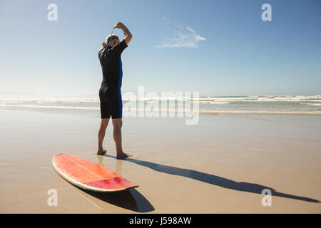 Heckansicht des senior Mann mit Neoprenanzug stehend von Surfbrett am Strand Stockfoto