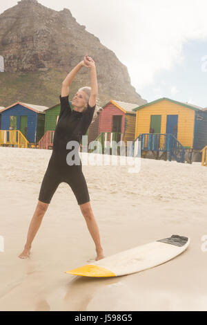 Lächelnde Frau ausdehnen, während sie von Surfbrett stehend gegen Hütten Stockfoto