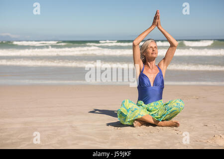 Ältere Frau, die Yoga praktizieren, während der Sitzung gegen klaren Himmel am Strand Stockfoto