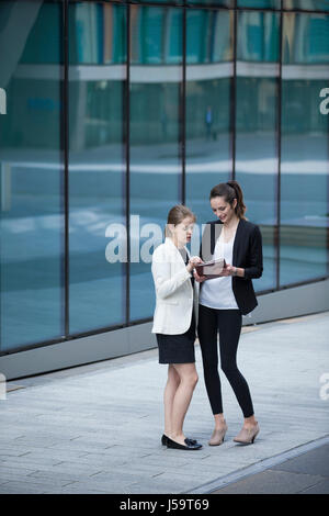 Zwei kaukasischen Geschäftsfrauen mit digitalen Tablettcomputer. Bild in voller Länge. Draußen, ein modernes Bürogebäude. Stockfoto