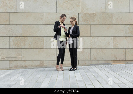 Zwei kaukasischen Geschäftsfrauen mit einem Smartphone, außerhalb, an eine Wand gelehnt. Stockfoto