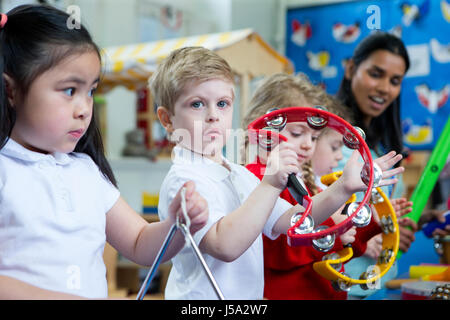 Kindergarten-Kinder spielen mit Musikinstrumenten im Klassenzimmer. Ein kleiner Junge schaut die Kamera mit einem Tamburin. Stockfoto