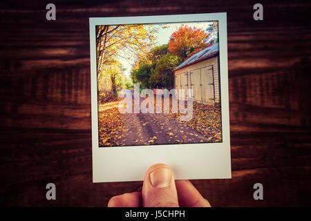 Männliche Hand, die Polaroid-Foto von Landstraße vorbei an hölzernen Schuppen zwischen gelb und orange Bäumen im Herbst Australien. Reise-Erinnerungen-scrapbooking Stockfoto