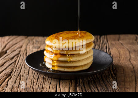 Ahornsirup auf einen Stapel von frischen Pfannkuchen, sitzt auf einem rustikalen Holztisch gegossen wird. Stockfoto