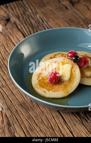 Drei frische Pfannkuchen beträufelt mit Sirup und Beeren in einer blauen Schüssel sitzt auf einem rustikalen Holztisch. Stockfoto