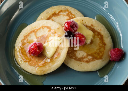 Drei frische Pfannkuchen beträufelt mit Sirup und Beeren in einer blauen Schüssel sitzt auf einem rustikalen Holztisch. Stockfoto