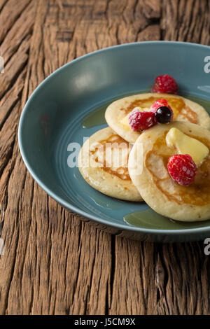 Drei frische Pfannkuchen beträufelt mit Sirup und Beeren in einer blauen Schüssel sitzt auf einem rustikalen Holztisch. Stockfoto