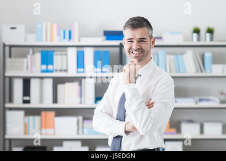 Zuversichtlich Geschäftsmann in seinem Büro stehen und blickte in die Kamera, er lächelt mit Hand am Kinn Stockfoto