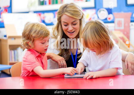 Weibliche Kindergärtnerin mit einem digitalen Tablet mit zwei Schülern in der Klasse. Stockfoto