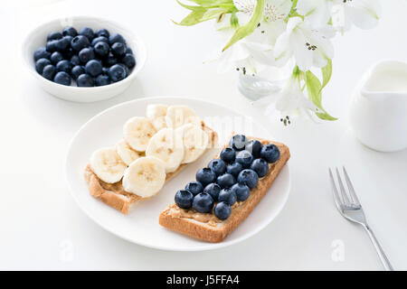 Gesunder Snack, gesundes Frühstück: zwei Toast mit Erdnussbutter zu verbreiten, Blaubeeren und Bananen auf weißen Tisch Stockfoto