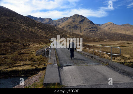 Einsame weibliche Hillwalker zu Fuß auf der Beton-Brücke über den Fluss Cluanie mit der schottischen Berge Munro Aonach Luft Chritih in Glen Shiel, Stockfoto