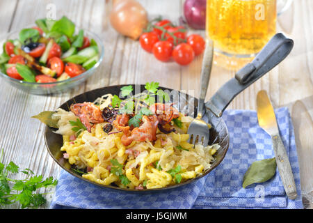 Schwäbische Spätzle mit Sauerkraut, gebratenem Speck und Zwiebelringe auf einem Holztisch mit einem Münchner Bier und einem Beilagensalat Stockfoto