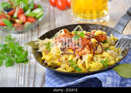 Schwäbische Spätzle mit Sauerkraut, gebratenem Speck und Zwiebelringe auf einem Holztisch mit einem Münchner Bier und einem Beilagensalat Stockfoto