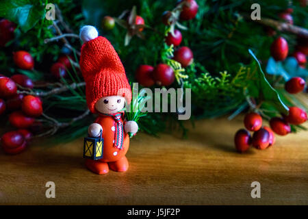 Kleine rote Holzfigur mit Bommel Mütze (Pudelmütze) und mit beleuchteter Laterne gegen grüne Weihnachtskranz mit roten Früchten Stockfoto