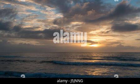 Sonnenaufgang hinter Wolken über den Strand mit Wellen im Vordergrund Stockfoto