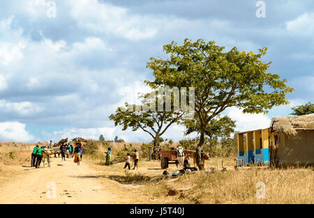 Eine unbefestigte Straße durch ein ländliches Dorf in Malawi, Afrika zeigt einen Ochsen Wagen Männer schieben Fahrräder und Brennholz auf ihren Köpfen tragen Frauen Stockfoto