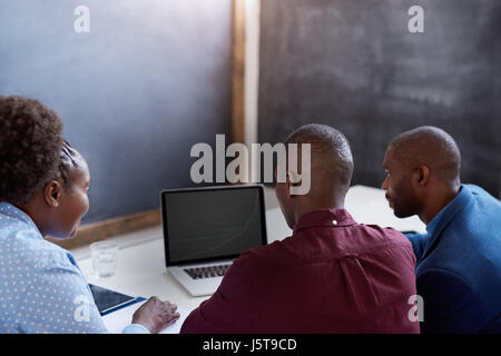 Rearview drei gekleidet beiläufig afrikanischen Kollegen sitzen zusammen an einem Tisch in einem modernen Büro an einem Notebook arbeitet Stockfoto