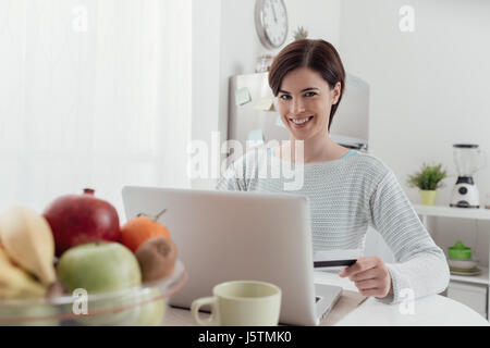 Lächelnde junge Frau mit einem Laptop in der Küche zu Hause verbinden, tut sie, Online-shopping und Kauf von Produkten unter Verwendung einer Kreditkarte Stockfoto