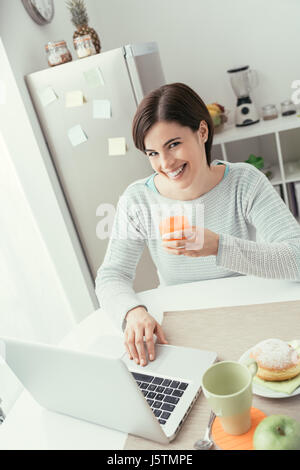 Junge, lächelnde Frau mit Frühstück in der Küche, ist sie Vernetzung mit einem Laptop und einen gesunden Orangensaft trinken Stockfoto
