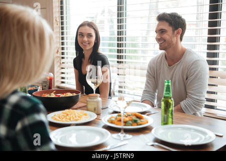 Fröhliche junge Menschen genießen Mahlzeit beim Sitzen am Esstisch in der Küche zusammen Stockfoto