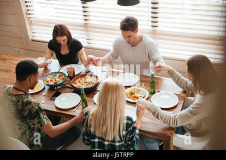 Gruppe von friedlichen Jugendlichen Hand in Hand und beten am Esstisch Stockfoto