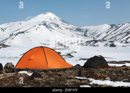 Outdoor-camping (touristische Zelt) auf Wiese, umgeben von Schnee in den Bergen im Hintergrund des Kegels der Awatscha-Vulkans auf Kamtschatka aktiv. Stockfoto