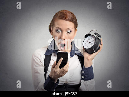 Überrascht, schockiert Geschäftsfrau mit Wecker, Smartphone mit lustige Gesichtsausdruck spät zur Besprechung isoliert graue Wand Hintergrund betrachten. H Stockfoto