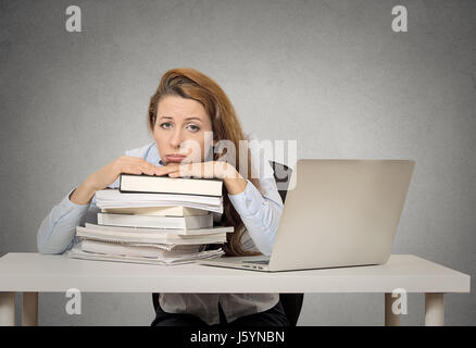 Demotiviert Studentin am Schreibtisch mit Haufen Bücher und Computer im sitzen gelangweilt lustige müde auf graue Wand Tafel Hintergrund isoliert. H Stockfoto