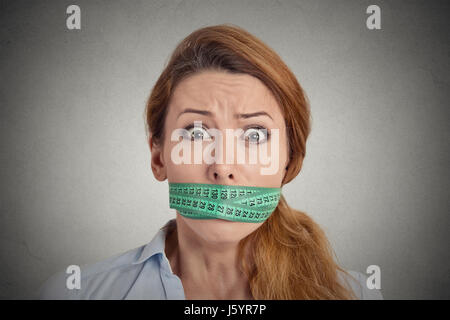 Porträt unglückliche junge Frau mit grünen Maßband den Mund isoliert graue Wand Background. Diät Lifestyle-Konzept Stockfoto