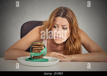 Porträt junge unglückliche Frau, die Lust auf Zucker süßes Gebäck aber besorgt über Gewichtszunahme, die am Tisch sitzen isoliert graue Wand Background. Menschliches Gesicht expr Stockfoto