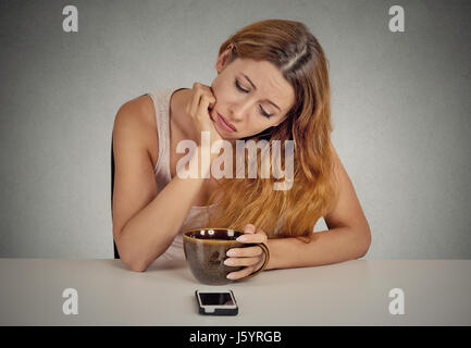 Traurig deprimiert junge Frau sitzt am Tisch Kaffee trinken Blick auf ihr Handy wartet auf einen Anruf SMS isoliert auf graue Wand hinter Stockfoto