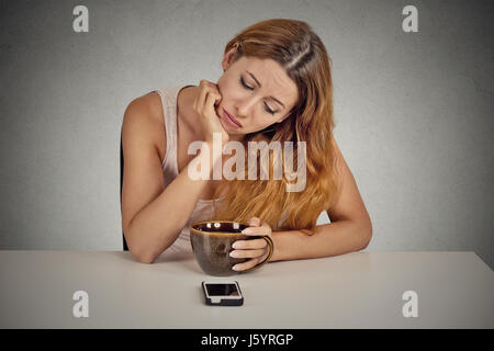 Traurig deprimiert junge Frau sitzt am Tisch Kaffee trinken Blick auf ihr Handy wartet auf einen Anruf SMS isoliert auf graue Wand hinter Stockfoto