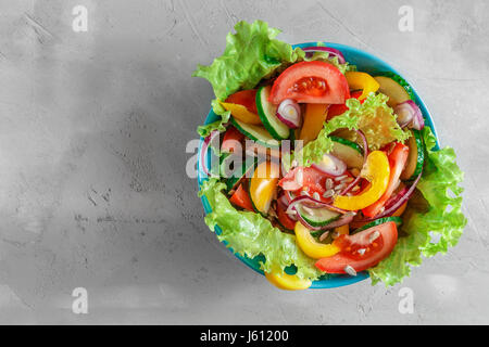 Frisches Gemüse Sommersalat mit Tomaten, Gurken, Paprika, Salat, Zwiebel und Sonnenblumenkerne in blau Schüssel über grauen Beton Hintergrund Stockfoto