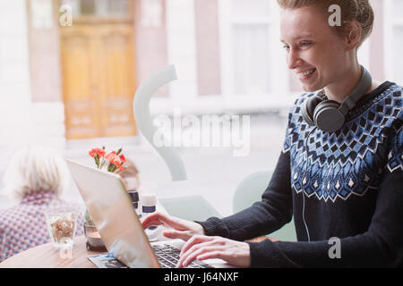 Lächelnde junge Frau mit Kopfhörern mit Laptop in Bürgersteig Café Fenster Stockfoto