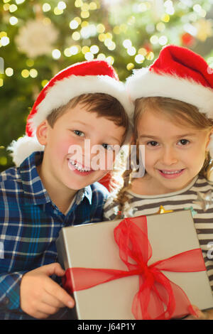 Porträt, Lächeln, Bruder und Schwester in Santa Hüte halten Weihnachtsgeschenk Stockfoto