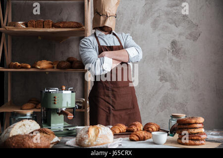 Bild des jungen Mannes Bäcker stehend mit Papiertüte auf Kopf mit Brille in Bäckerei in der Nähe von Brot. Stockfoto