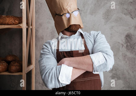 Bild des jungen Mannes Bäcker stehend mit Papiertüte auf Kopf mit Brille in Bäckerei in der Nähe von Brot. Stockfoto