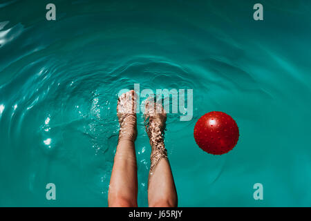 Eine Frau, tauchen ihre Füße in einem Swimmingpool, an einem warmen Sommertag Abkühlung. Eine rote Kugel schwebt in der Nähe von ihren Füßen, bewegt sich in die Wellen. Stockfoto