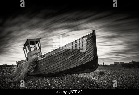Einzelne verlassene Fischerboot auf einem Kiesstrand mit langer Belichtung Wolken in Monochrom. Dungeness, England Stockfoto