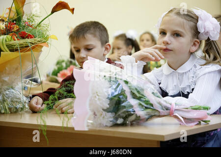 Der Beginn des akademischen Jahres. Elegante Kinder sitzen auf der Schulbank. Auf einer Schule Schreibtisch Blumen Lüge. Beide von ihnen zum ersten Mal sind in die Schule gekommen. Stockfoto