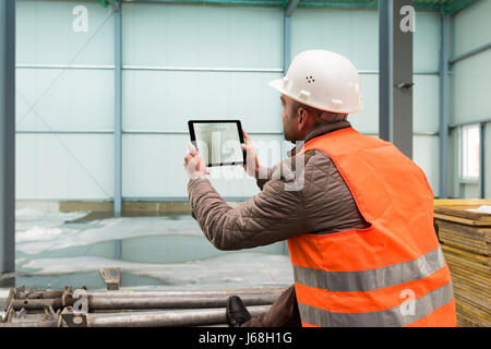 Bauleiter prüft das Innere eines neuen Lagers gebaut mit einem digitalen Tablet zeigt eine Karte in der Hand tragen eine Sicherheit Hel Stockfoto