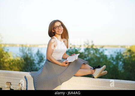 Junge attraktive Hipster Mädchen ist ein Buch zu lesen und Lächeln auf den Lippen. Sie ist in Gläser, Top, Rock und Turnschuhen bekleidet. Studentin ist Autodidakt. Studie Stockfoto