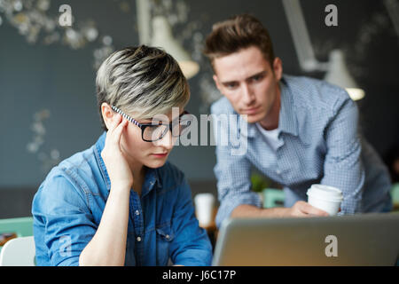 Porträt des jungen Geschäftsfrau trägt kreative Kurzhaarschnitt und Brille schauen verwirrt bei der Arbeit mit Laptop im Büro, Kollegen betrachten Stockfoto