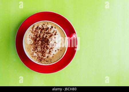 Blick von oben auf einer schaumigen Tasse Cappuccino Kaffee in eine rote Tasse und Untertasse und auf eine bunte, grüne Fläche. Stockfoto