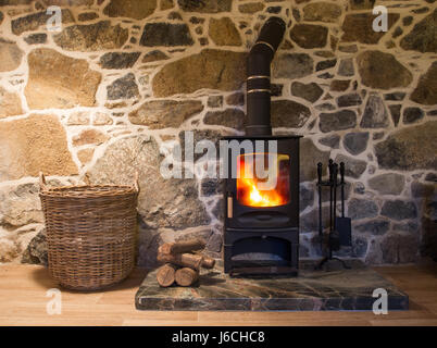 Das Innere eines gemütlichen Holzofen Landhaus aus Stein gemauerten zeigt eine Kamin-Szene mit einem hölzernen brennenden auf einem Herd mit Protokollen und einen Korb. Stockfoto