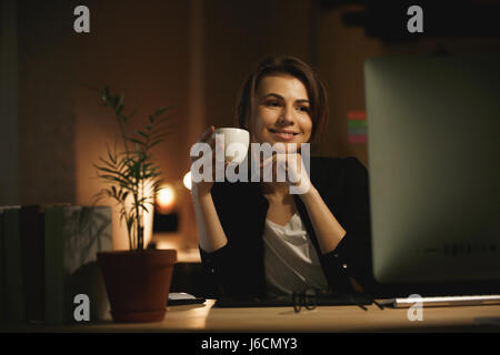 Foto der glückliche junge Frau Designer sitzen drinnen in der Nacht mit Computer und trinken Kaffee. Auf der Suche zur Seite. Stockfoto