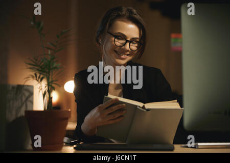 Bild der fröhliche junge Dame Designer sitzen drinnen in der Nacht mit Computer und Lesebuch. Auf der Suche zur Seite. Stockfoto