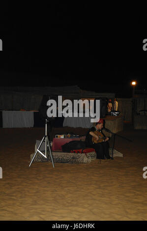 Nachtflaschenporträt arabischer Mann in schwarzer traditioneller Kleidung Gitarre spielt, Sofa auf Wüstensand sitzt, Alpha Tours Desert Camp, Dubai, VAE Stockfoto